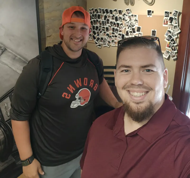 Seth Scrimo and Chad Tennent smiling in a coffee shop in front of a wall of polaroid photos